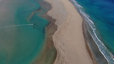 Playa-De-La-Barca,-Fuerteventura:-Vista-Aérea-Viajando-Sobre-La-Hermosa-Playa-Y-Una-Persona-Practicando-Windsurf