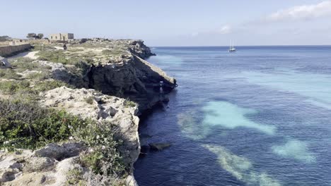 Steep-coastal-sea-cliff-drops-off-into-turquoise-blue-ocean-water-with-sailboat