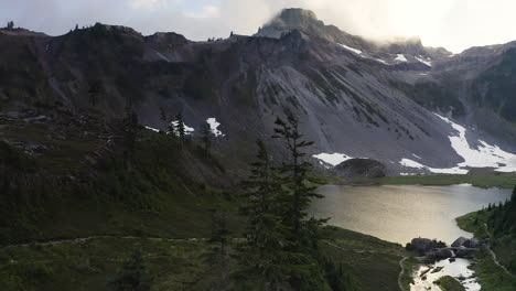 Drone-rises-above-wispy-evergreen-trees-to-alpine-lake-at-base-of-avalanche-path-at-sunset