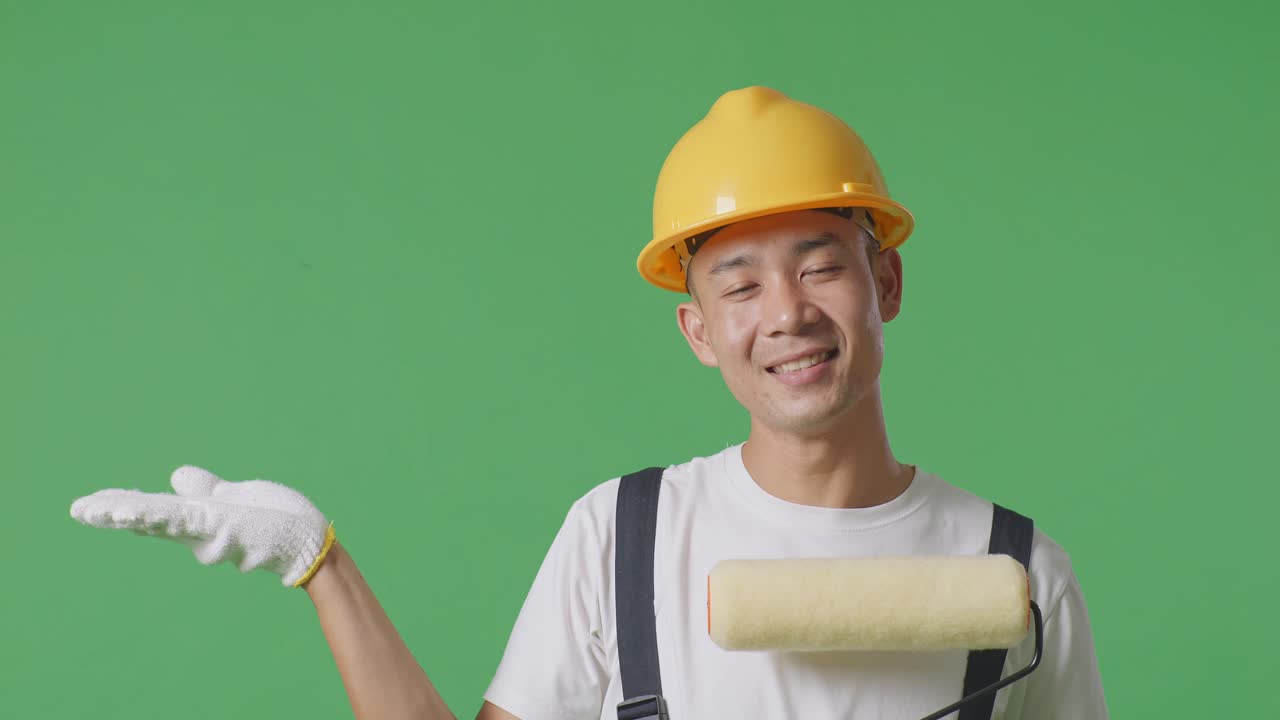 Close Up Of Asian Man Painter Wearing Safety Helmet Smiling And Pointing To Side While Standing In The Green Screen Background Studio