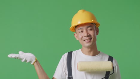 close up of asian man painter wearing safety helmet smiling and pointing to side while standing in the green screen background studio