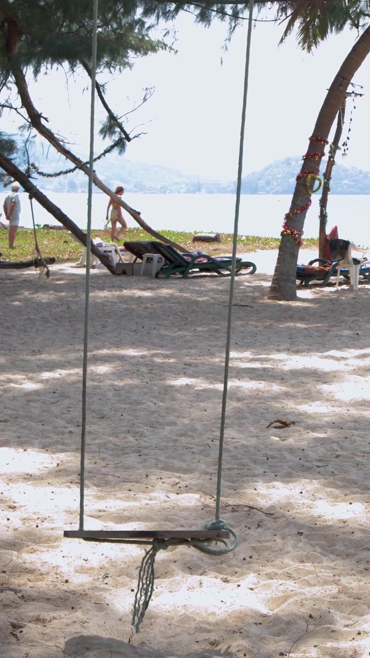 En statisk vidvinkelbild fångar en tom trärgunga som svajar under träd på Phukets strand.
