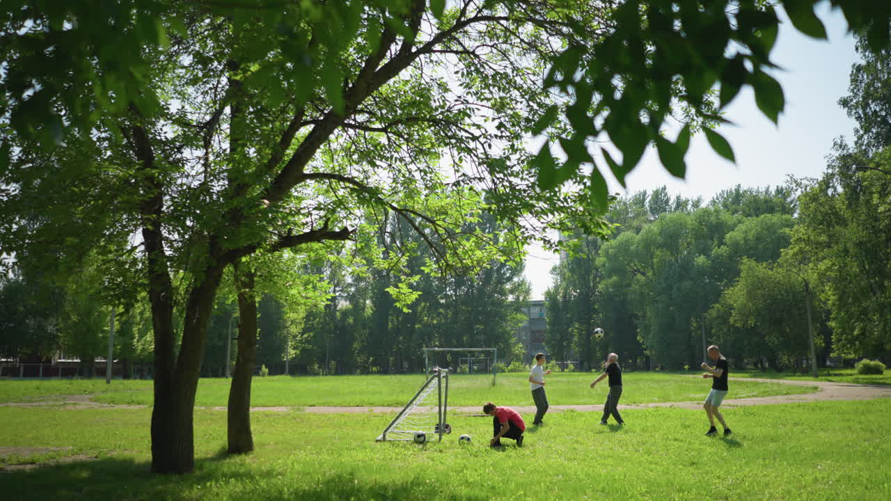 una familia pasa un día divertido al aire libre, el niño de la camiseta roja ata sus cordones cerca de un poste de la portería, mientras el abuelo y el nieto juegan al fútbol y el otro hombre hace ejercicio