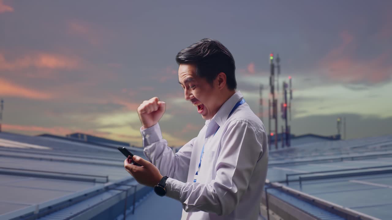 ภาพด้านข้างของพนักงานชายชาวเอเชียมืออาชีพพร้อมสมาร์ทโฟนของเขา กำลังตรวจสอบแผงโซลาร์เซลล์บนหลังคาโรงงานอุตสาหกรรม เขายกกำปั้นขึ้นด้วยความยินดีหลังจากตรวจสอบข้อมูลบนสมาร์ทโฟน
