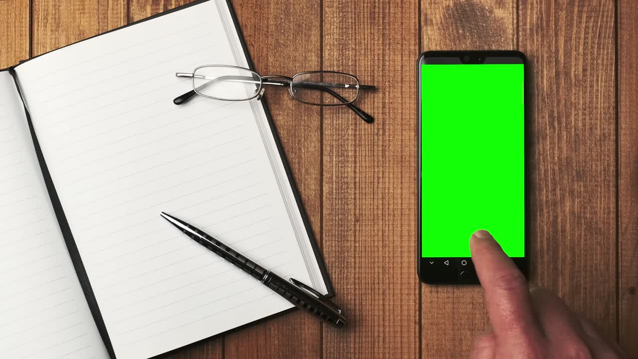 Top view of an open notebook with blank pages, glasses and a cell phone with a green screen on a wooden table. Mobile app mockup.
