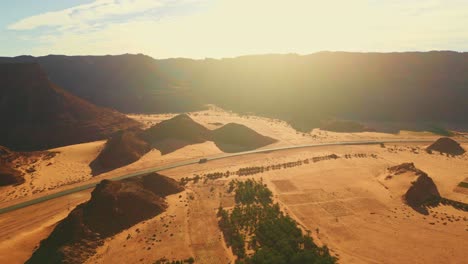 Autobahn-Mit-Wenig-Verkehr-Auf-Asphaltstraße-Durch-Wüste-Mit-Grünen-Palmen,-Canyon-Mit-Orangefarbenem-Felsberg-Und-Gelbem-Sand-Bei-Sonnenuntergang