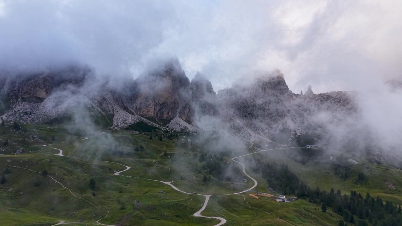 Vista aerea con drone di strade di montagna tortuose che attraversano i verdi pendii del Passo Gardena sotto le aspre cime delle Dolomiti parzialmente coperte da nebbia e nuvole basse