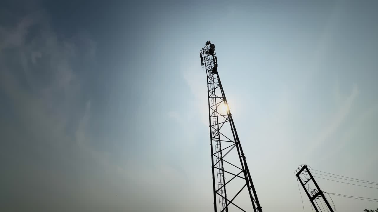 Scatto grandangolare di una torre di comunicazione mobile che si erge contro un cielo sereno, con la luce del sole che filtra attraverso il traliccio d'acciaio, a simboleggiare connettività, tecnologia e infrastruttura