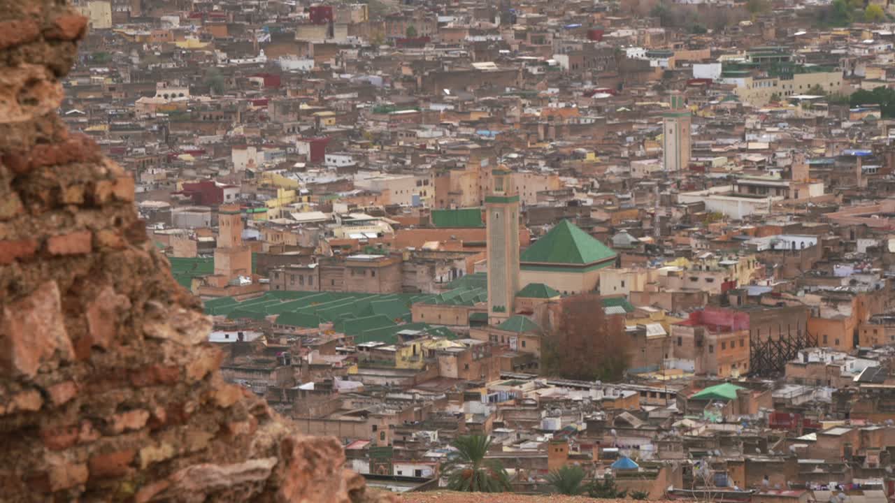 Una vista panoramica aerea cinematografica dall'alto che guarda attraverso un'antica muraglia in terra battuta, catturando la densa architettura storica e i minareti con tegole verdi della città murata di Fez.