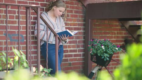 niña lee un libro mientras se apoya contra una pared de ladrillos y escaleras al lado de una bicicleta decorada con flores