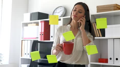 businesswoman standing near glass wall with sticky notes and using smartphone