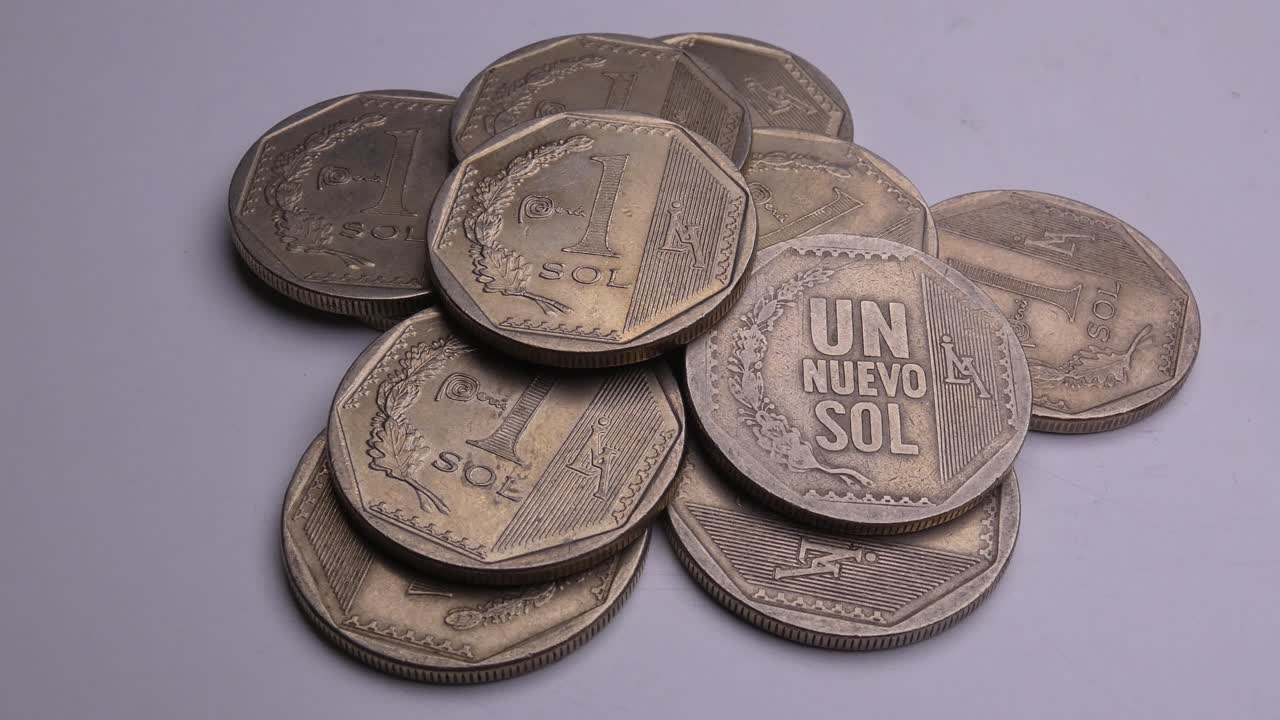Many silver colored coins in a white background with a light above. The table moves and slowly puts the coins into frame. The coins are PEN or Peruvian soles, the currency from Peru