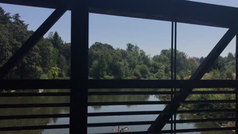 Handheld-footage-through-the-rusted-railing-of-a-footbridge-over-a-calm-and-slow-moving-river-during-the-summer-in-Oregon