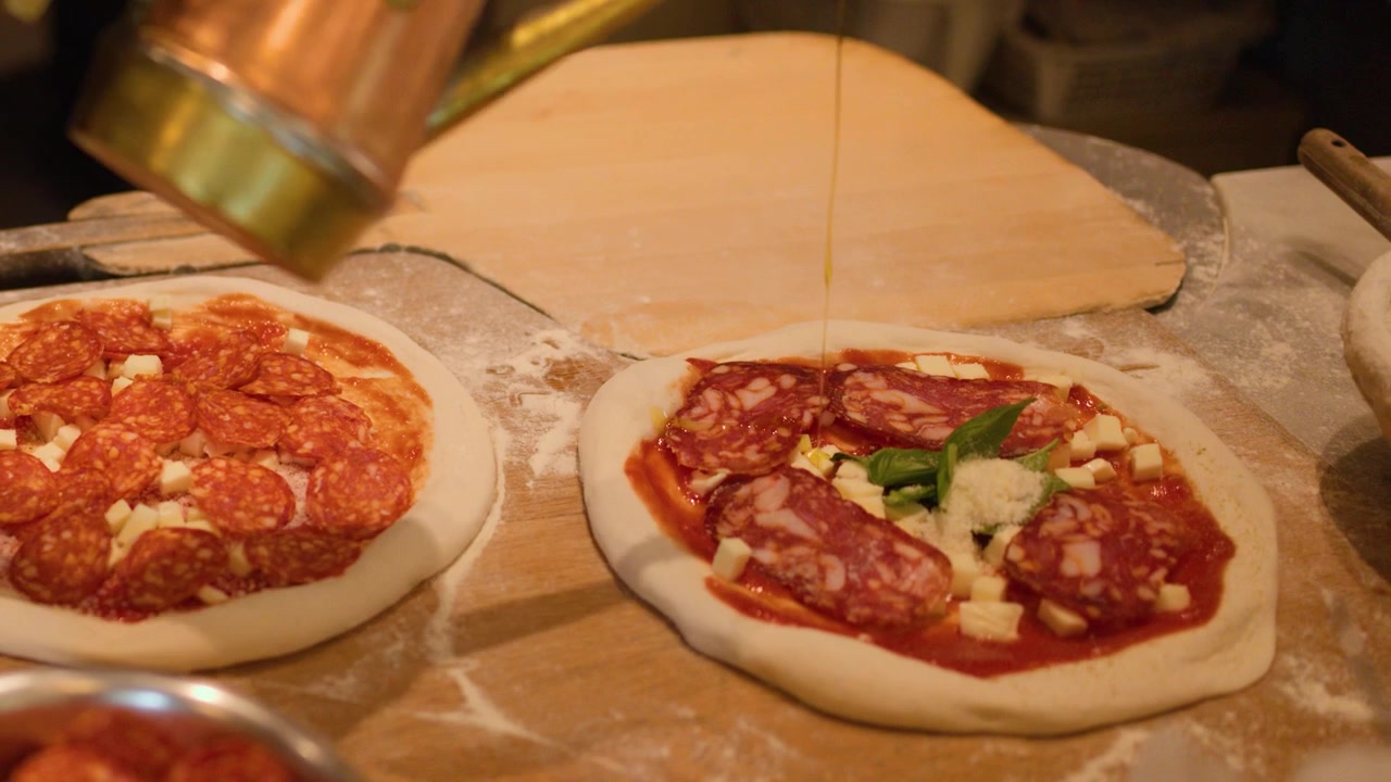 Primo piano di uno chef che aggiunge condimenti e olio d'oliva a pizze crude su un bancone di legno.
