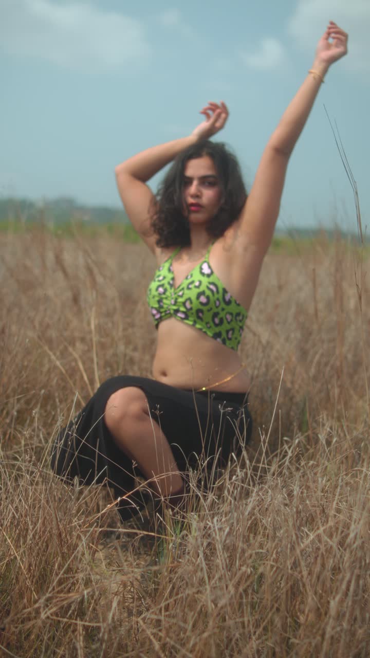 Young woman poses in dry grass field, arms raised, wearing patterned top, cloudy sky