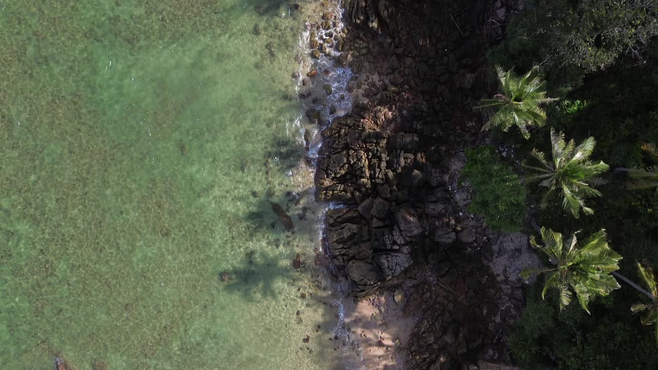 Top down drone footage of a tropical shoreline in Koh Lanta, Thailand, showing clear turquoise sea, palm trees and rocky beach formations in a calm and scenic island landscape