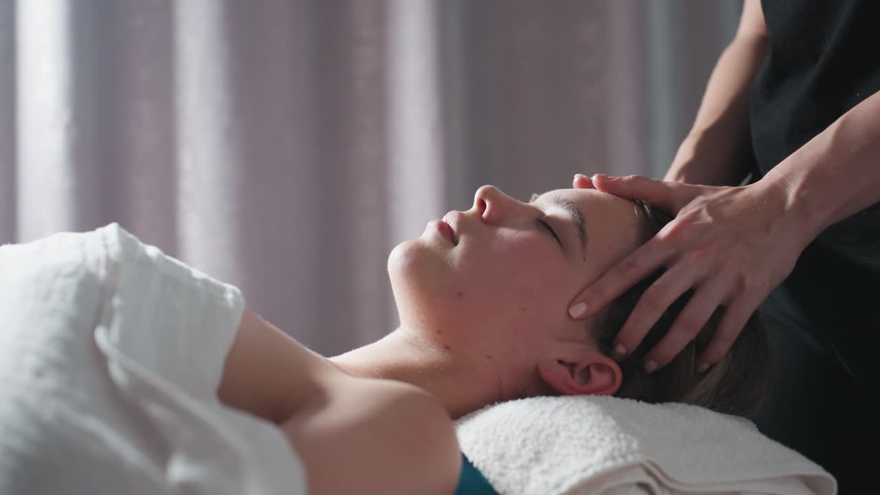 Calm scene of woman resting on folded towel while therapist gently massages forehead and scalp, soft natural light filtering through gray curtain, creating peaceful and relaxing spa environment