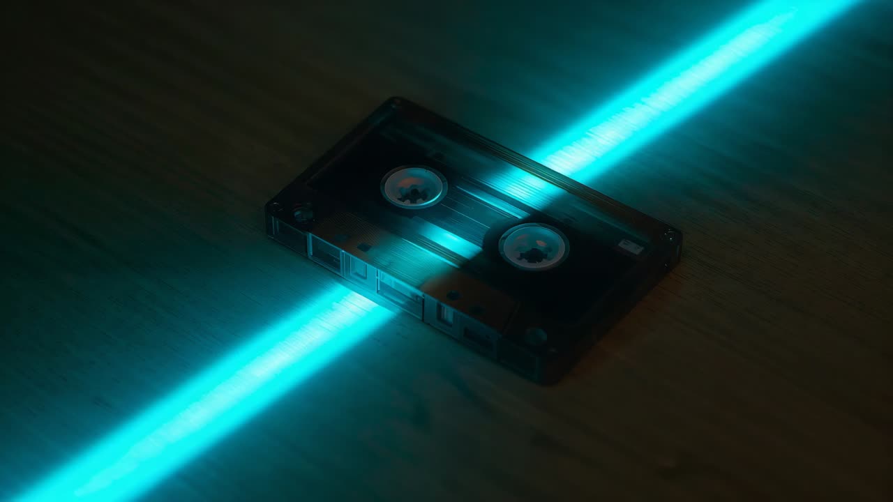 Sweeping cyan beam sliding across wooden tabletop, lighting clear cassette tape and rotating reels