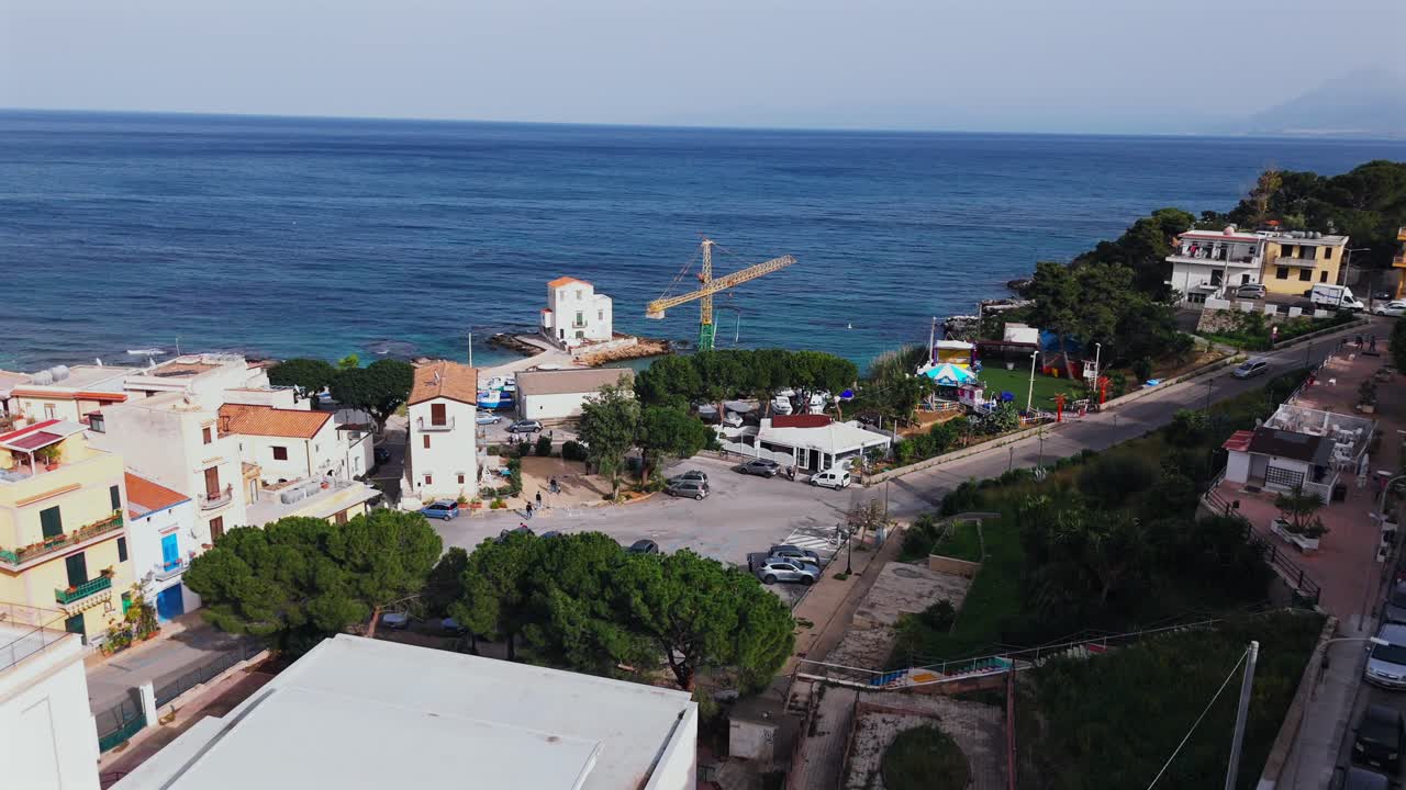 Slow motion aerial footage over Porticello’s coastal edge in Sicily showing harbor structures, a construction crane and waterfront activity near the Tyrrhenian Sea with distant mountains