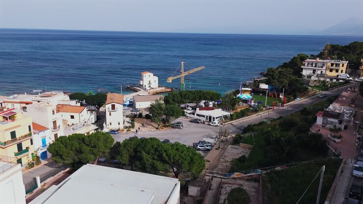 Filmagem aérea em câmera lenta sobre a orla costeira de Porticello na Sicília, mostrando estruturas portuárias, um guindaste de construção e atividade à beira-mar perto do Mar Tirreno com montanhas distantes.