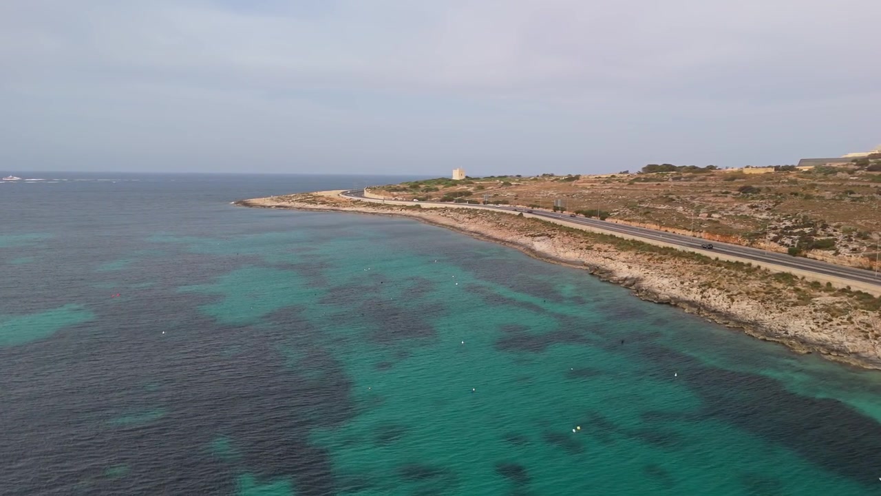 A Salina, sulla costa maltese, una strada costiera con veicoli in movimento corre lungo la costa rocciosa e le limpide acque del Mediterraneo sotto un cielo diurno nuvoloso, ripreso da drone.
