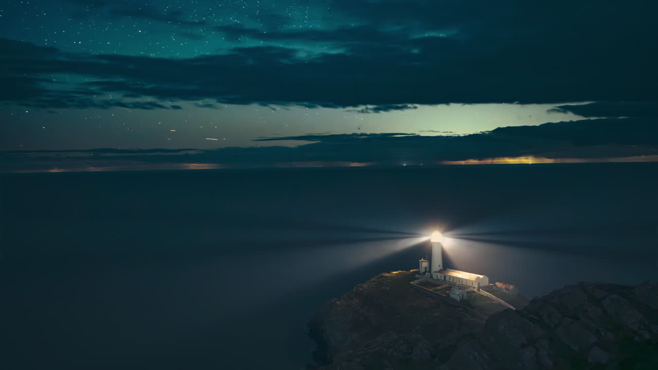 Timelapse of South Stack Lighthouse, Anglesey. A rotating beam cuts through cloudy night sky, stars, and sea. A stunning and atmospheric coastal scene of a Welsh maritime landmark