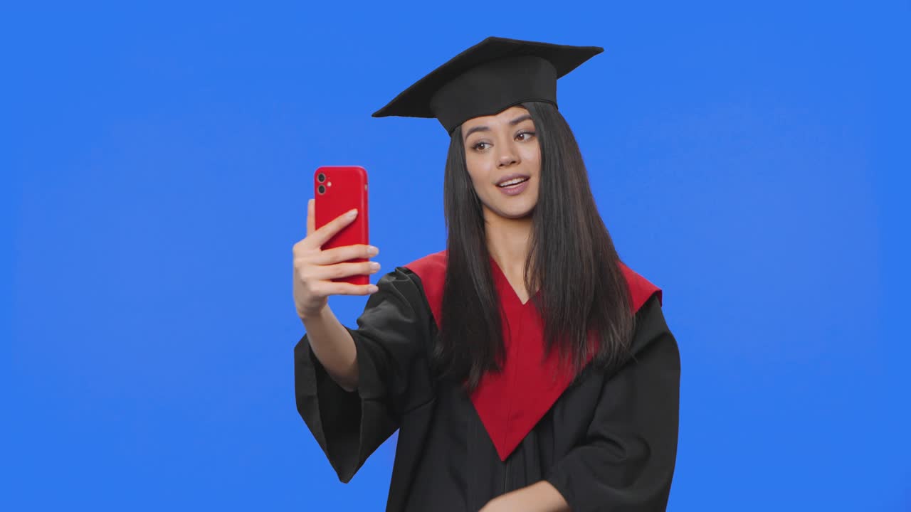 Portrait of female student in graduation costume, holding diploma and talking on video call using phone. Young woman posing in studio with blue screen background. Close up. Slow motion ready 59.94fps