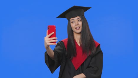 retrato de una estudiante en disfraz de graduación, sosteniendo un diploma y hablando en una llamada de video usando el teléfono. mujer joven posando en el estudio con fondo de pantalla azul. primer plano. cámara lenta lista 59.94fps