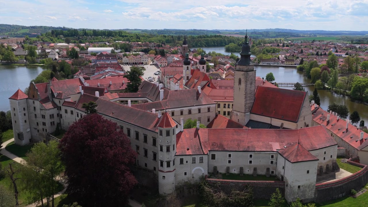 Vista aérea de drone de torres gêmeas de igreja barroca e uma torre do relógio medieval elevando-se acima de edifícios com telhados vermelhos, com um lago e colinas arborizadas ao fundo, Telč, República Tcheca