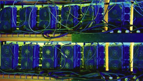 multiple asic bitcoin miners with prominent cooling fans and tangled wiring mounted on metal shelves in a server rack. close up pan right