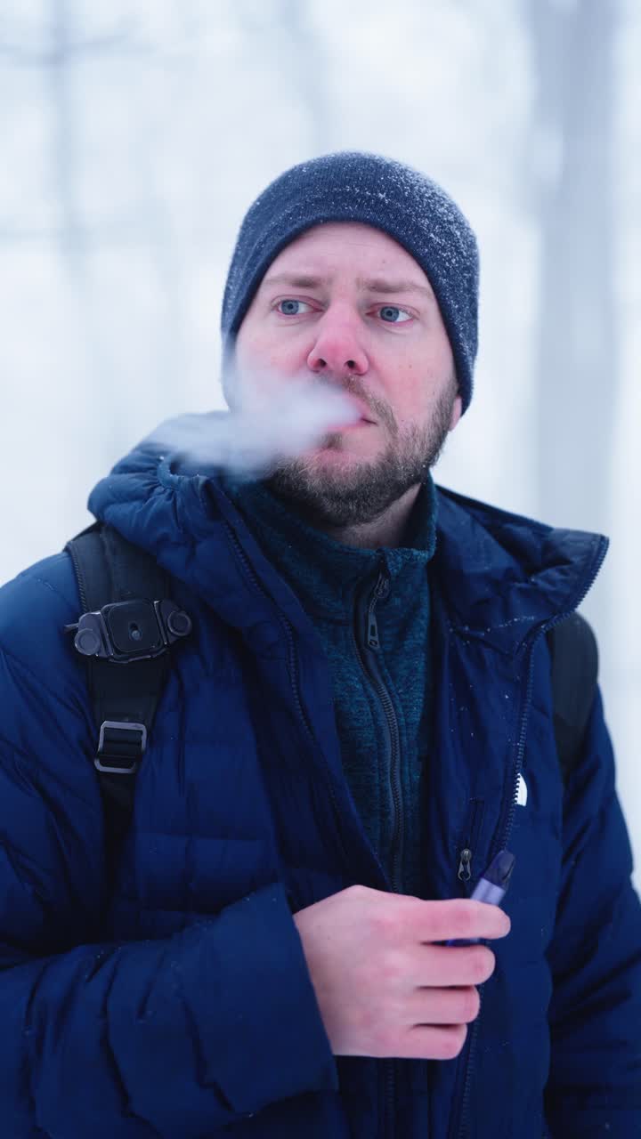 Nærbillede af mand der damper e-cigaret udendørs om vinteren. Sne på hat og kold ånde skaber et stemningsfuldt sæsonportræt i en tåget skovomgivelse
