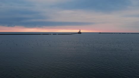 aerial flying low above water lake michigan towards milwaukee breakwater light, captured at sunrise, historical landmark in wisconsin, cinematic shot with birds on lake