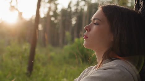 una mujer cansada se apoya contra un árbol en el bosque al atardecer. una joven siente emociones negativas después de una ruptura difícil encontrando consuelo del estrés en la naturaleza