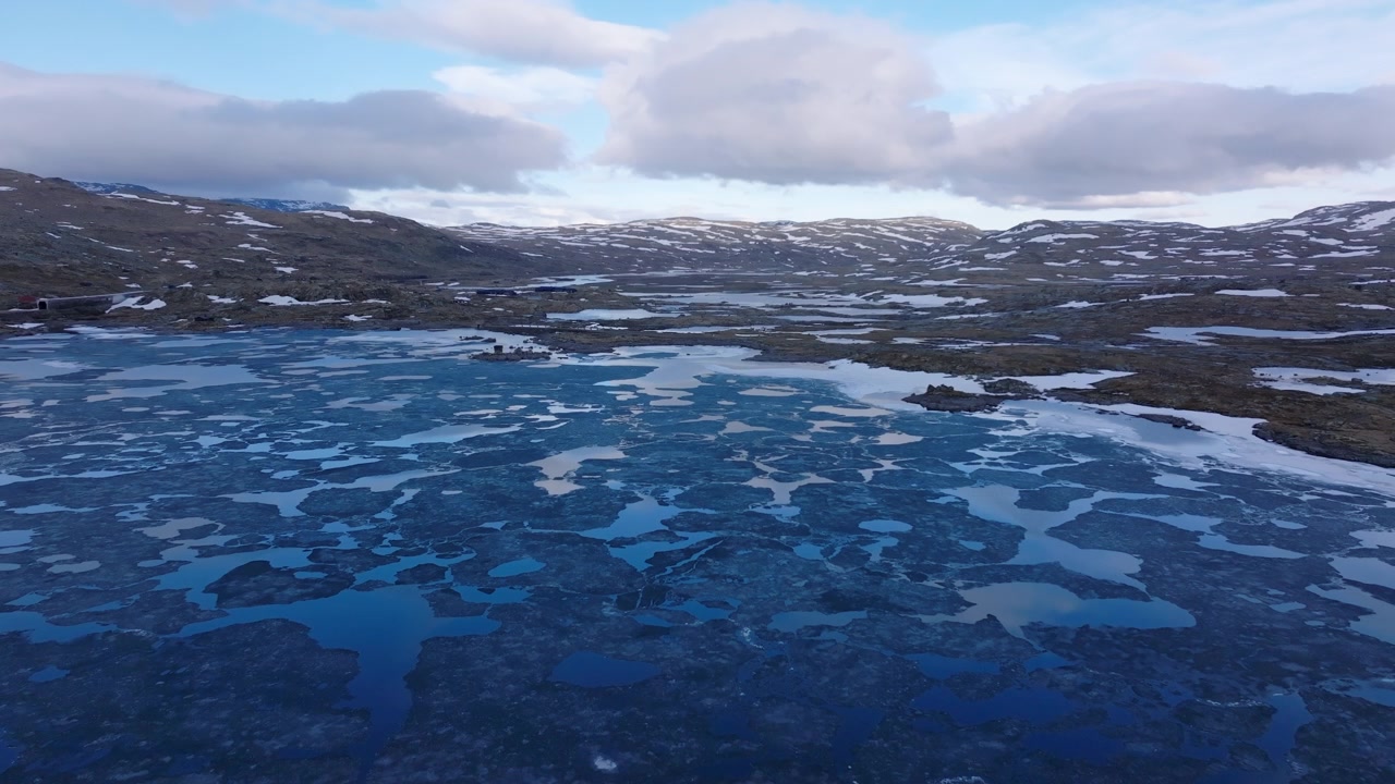 Droneoptagelser over en levende blå bjergsø med flydende is og spejllignende skyrefleksioner i det norske højland