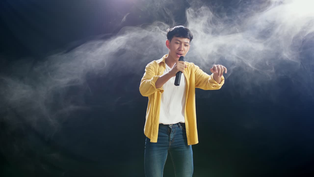 마이크를 들고 있는 젊은 아시아 소년이 색 연기가 있는 검은색 배경에서 을 치고 있다.