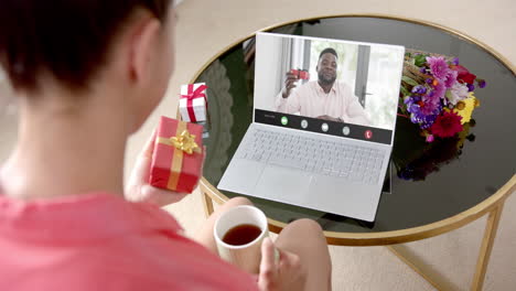 caucasian woman holding red gift using laptop with african american man on screen