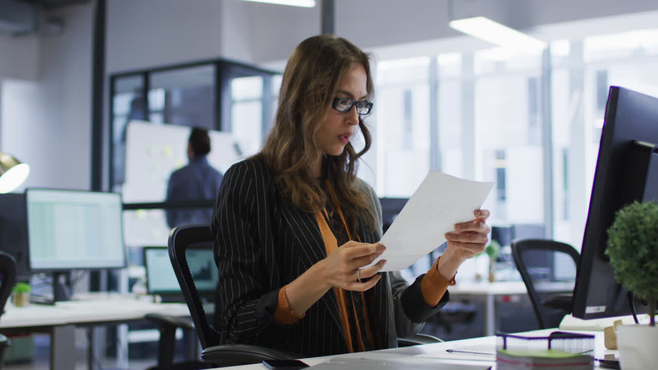 mujer de negocios caucásica sentada en el escritorio leyendo un documento y quitándose las gafas en la oficina