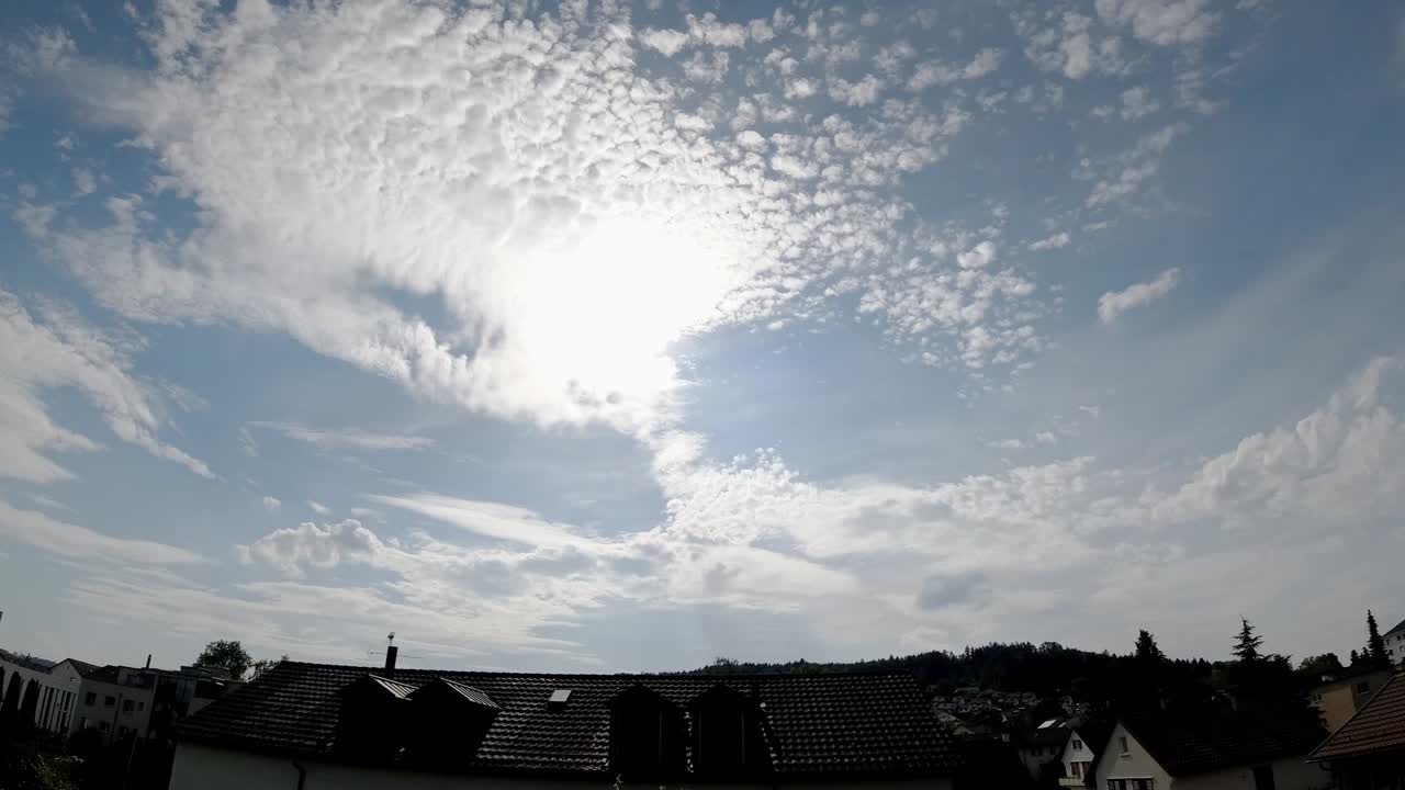 Time-lapse of the clouds and the sun setting in a blue sky with some houses in the landscape.