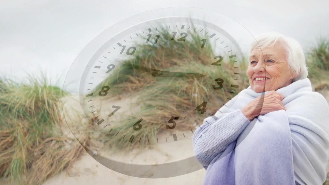 Senior woman feeling cool breeze, wrapping shawl on dune, clock overlay covering subject, wellness