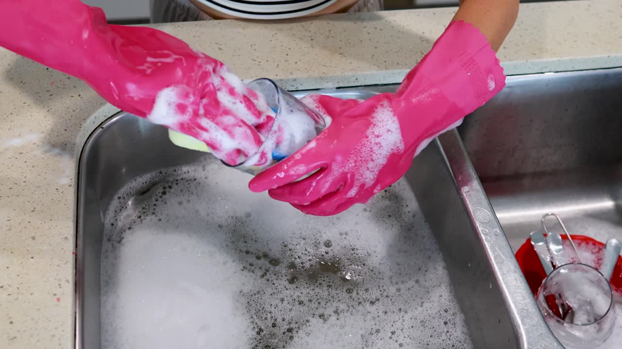 A person wearing pink gloves cleans a glass with a sponge in a kitchen sink filled with soapy water