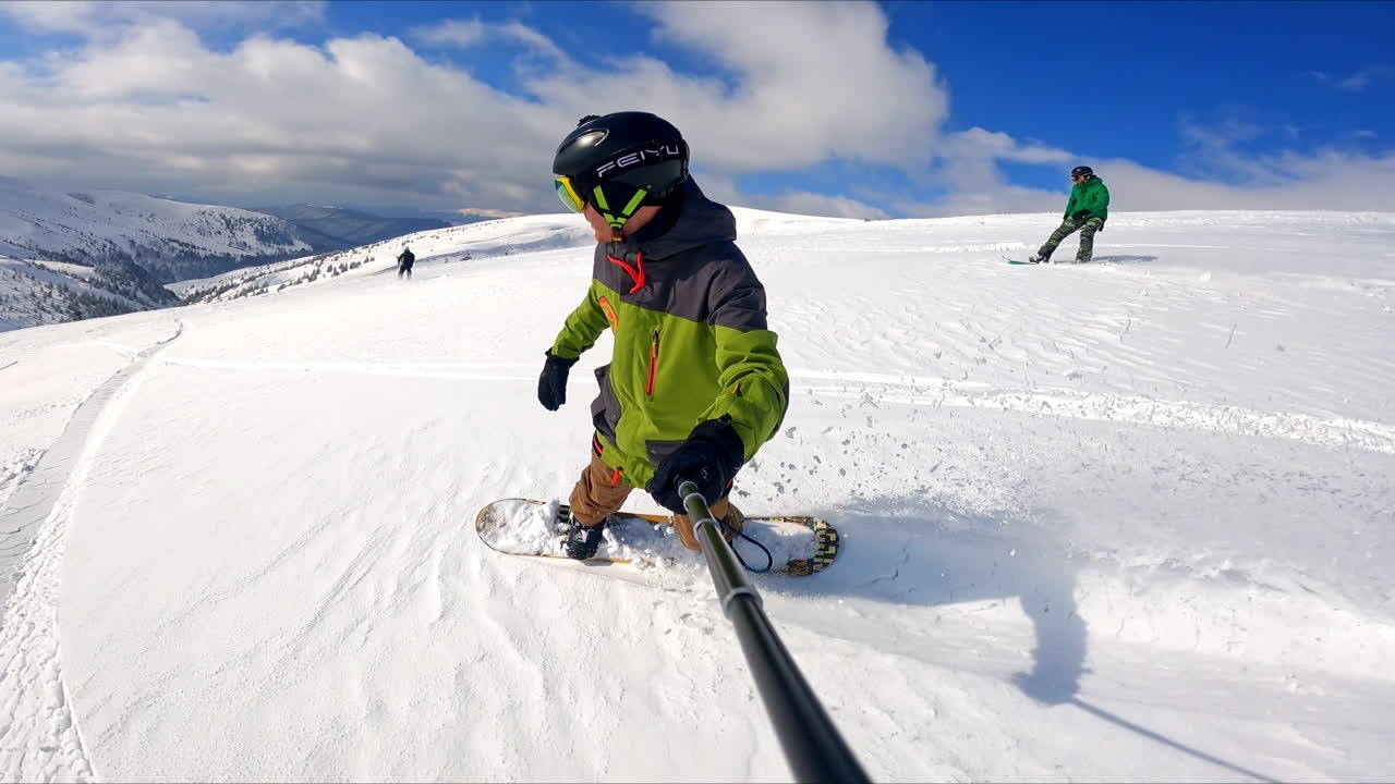 Male snowboarder taking selfie video of his ride. Two other sportsmen ride down the mountain at backdrop.