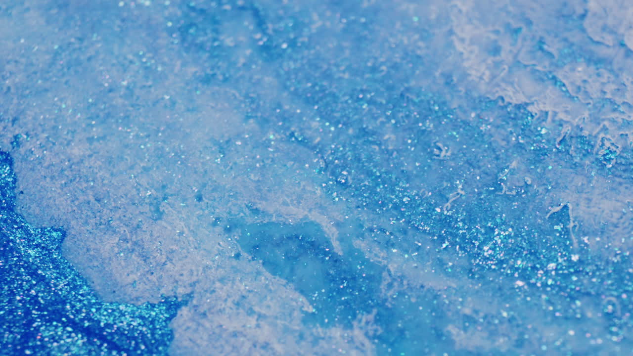 Macro shot of blue glitter ink reacting to injected air, creating wave-like ripples and shimmering motion in the fluid surface