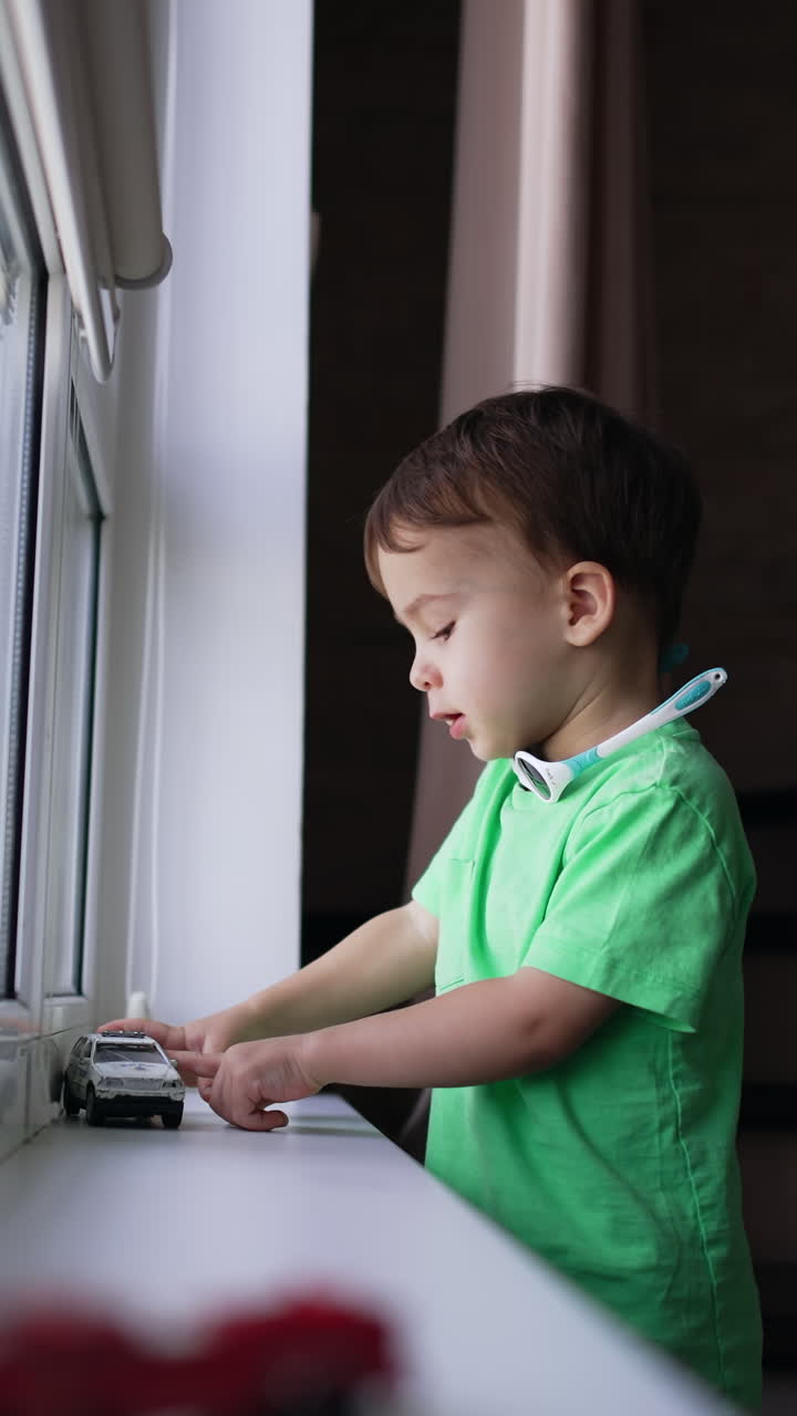 Caucasian baby in green t-shirt playing at home. Focused kid pushing a toy car by the window-sill. Vertical video