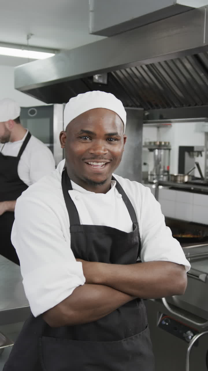 Happy african american male chef standing with arms crossed in kitchen, slow motion, vertical