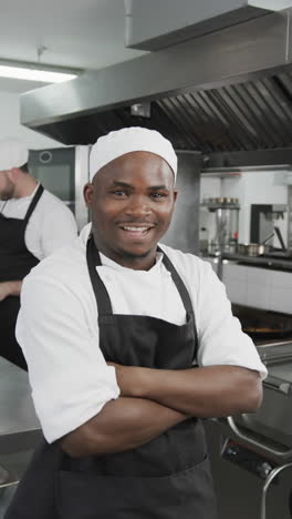 happy african american male chef standing with arms crossed in kitchen, slow motion, vertical