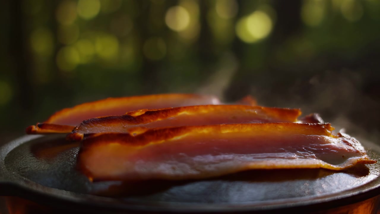 Vista ravvicinata dal basso di striscioline di pancetta che friggono in una padella di ghisa calda, con vapore visibile, bordi che si arricciano, grasso sciolto, superficie lucida e un graduale passaggio dal rosa al marrone dorato, con pancetta, maiale, carne.