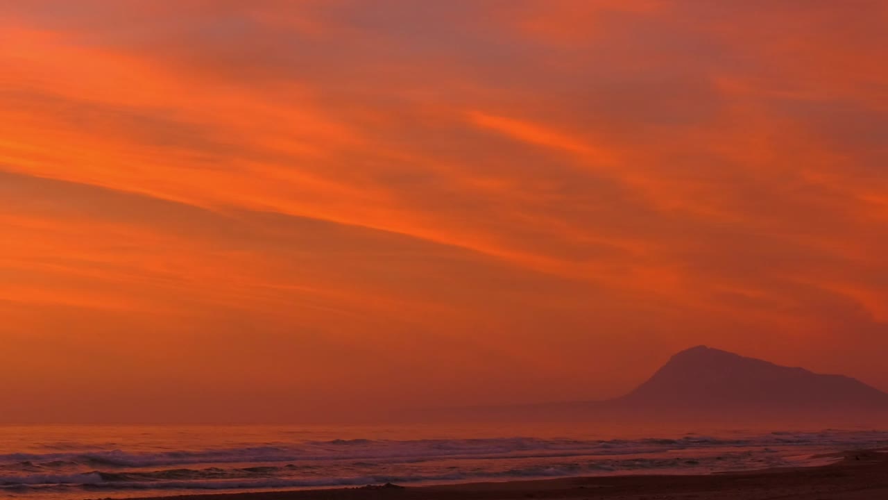 Orange dawn skies over beach with rolling waves and mountain in background, panning shot