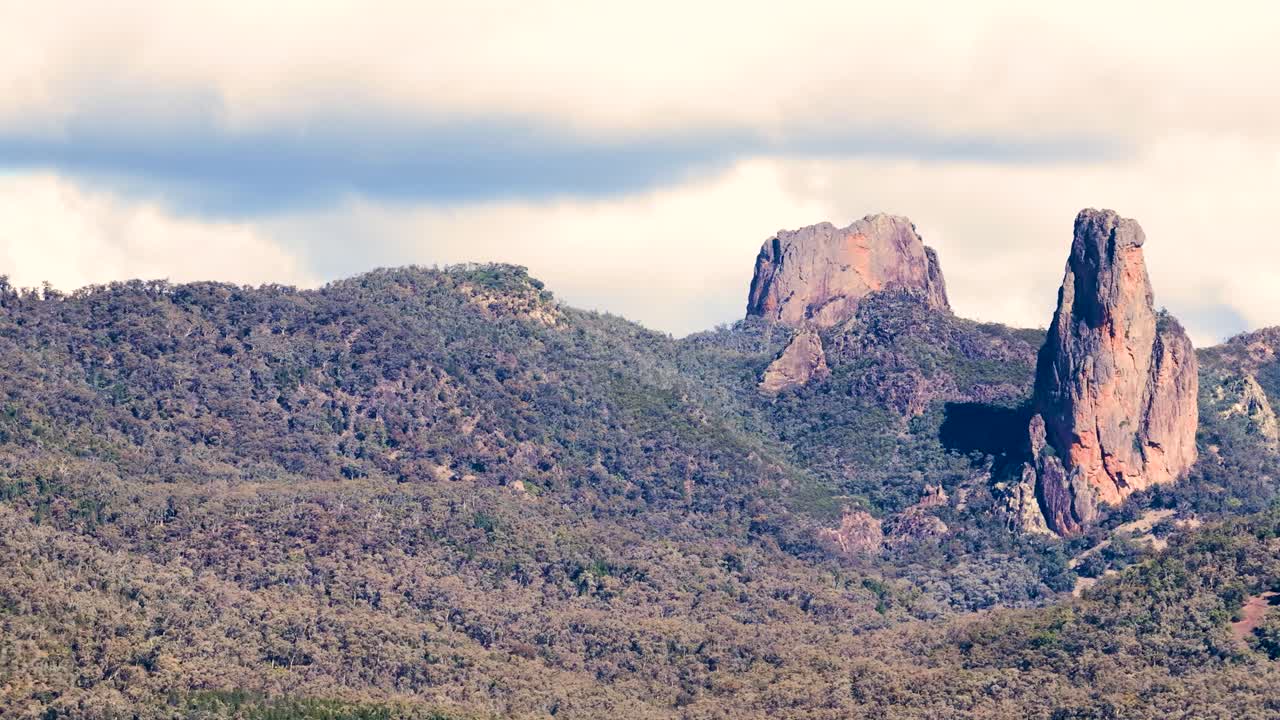 Wide timelapse pan across rugged Siding Spring mountains and forested hills under soft daylight, revealing dramatic rock formations and partly cloudy sky