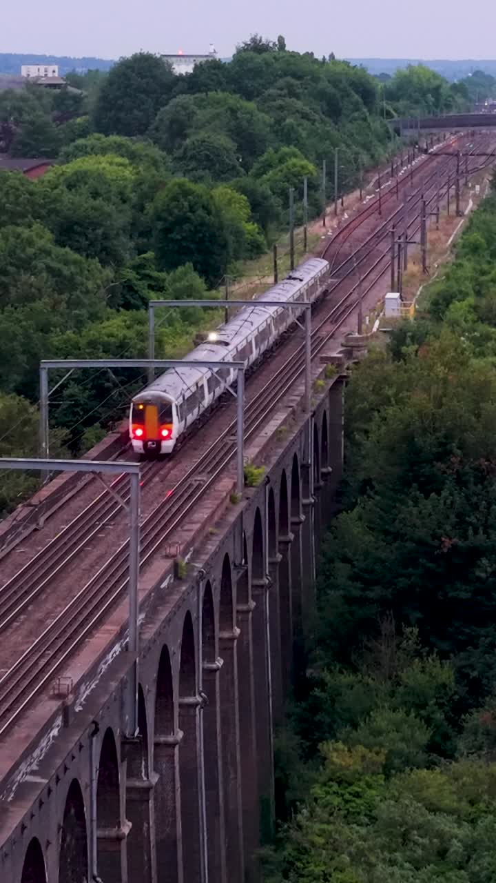 Vertical drone shot reversing along railway track at Digswell Viaduct as a train moves forward at dusk. The viaduct is framed by green trees, houses and soft early evening light