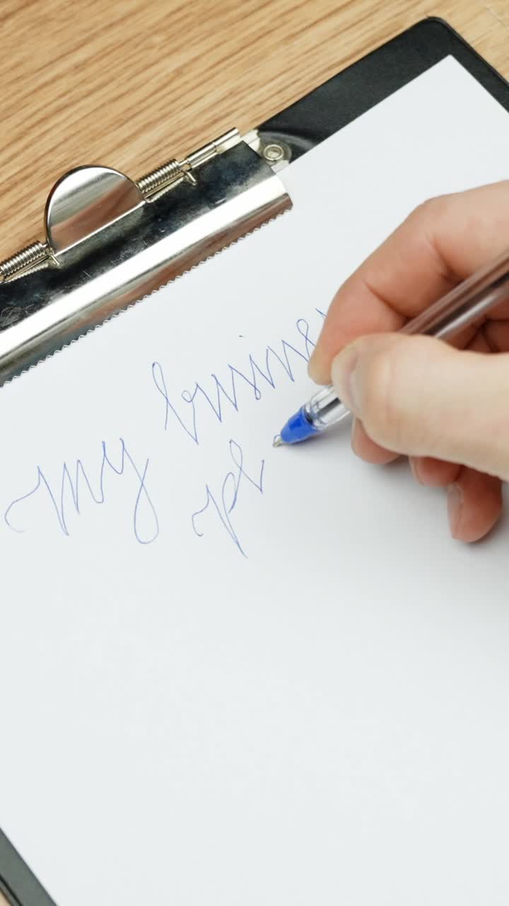 Close-up of a hand using a blue pen to write "my business plan" in cursive on a clipboard in Vertical composition emphasizing focus and precision during early planning steps on a wood desk surface.